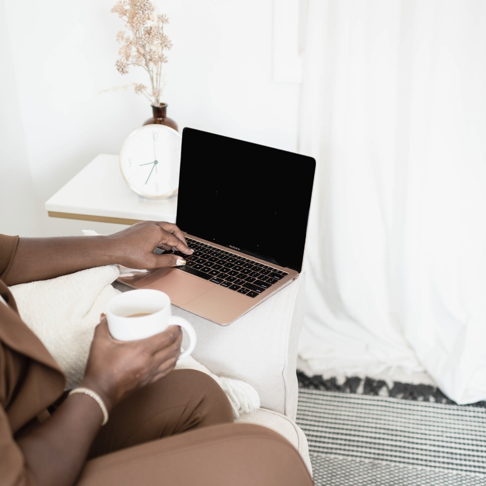 business woman with brown suite typing on rose gold laptop and holding cup coffee